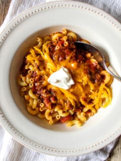 Bowl of chili over macaroni with toppings.