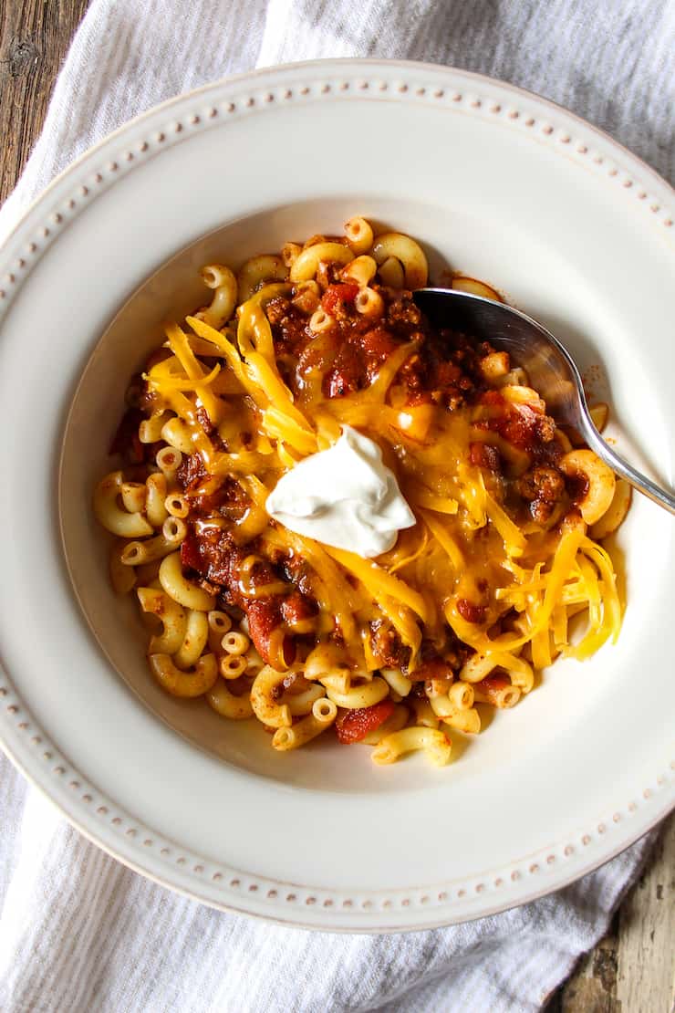 Bowl of chili over macaroni with toppings.
