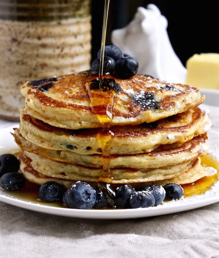 Cornmeal Blueberry pancakes, side photo of stack of pancakes with syrup being poured over them.