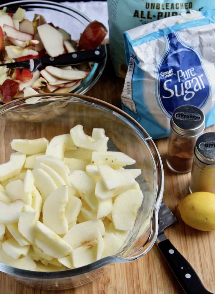 Sliced apples in bowl surrounded by ingredients for pie.