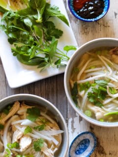 Two bowls of chicken pho with herbs and hoisin sauce.