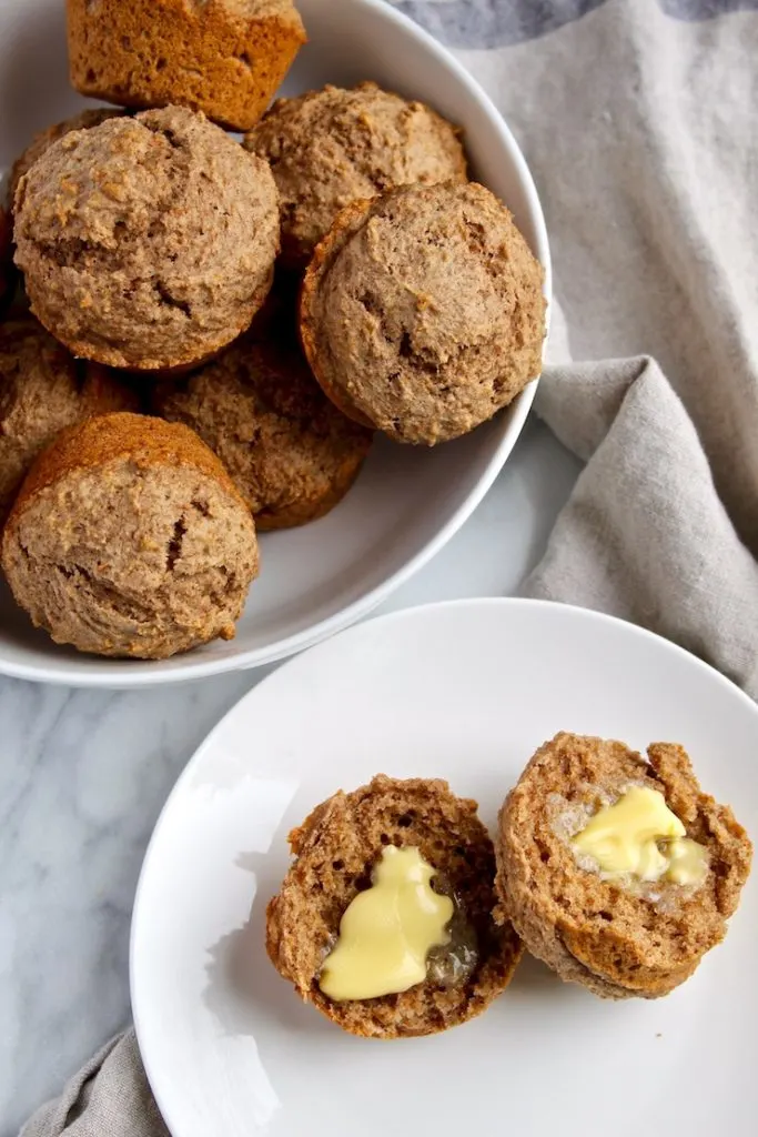 Muffin split with butter on plate, bowl of muffins in background.