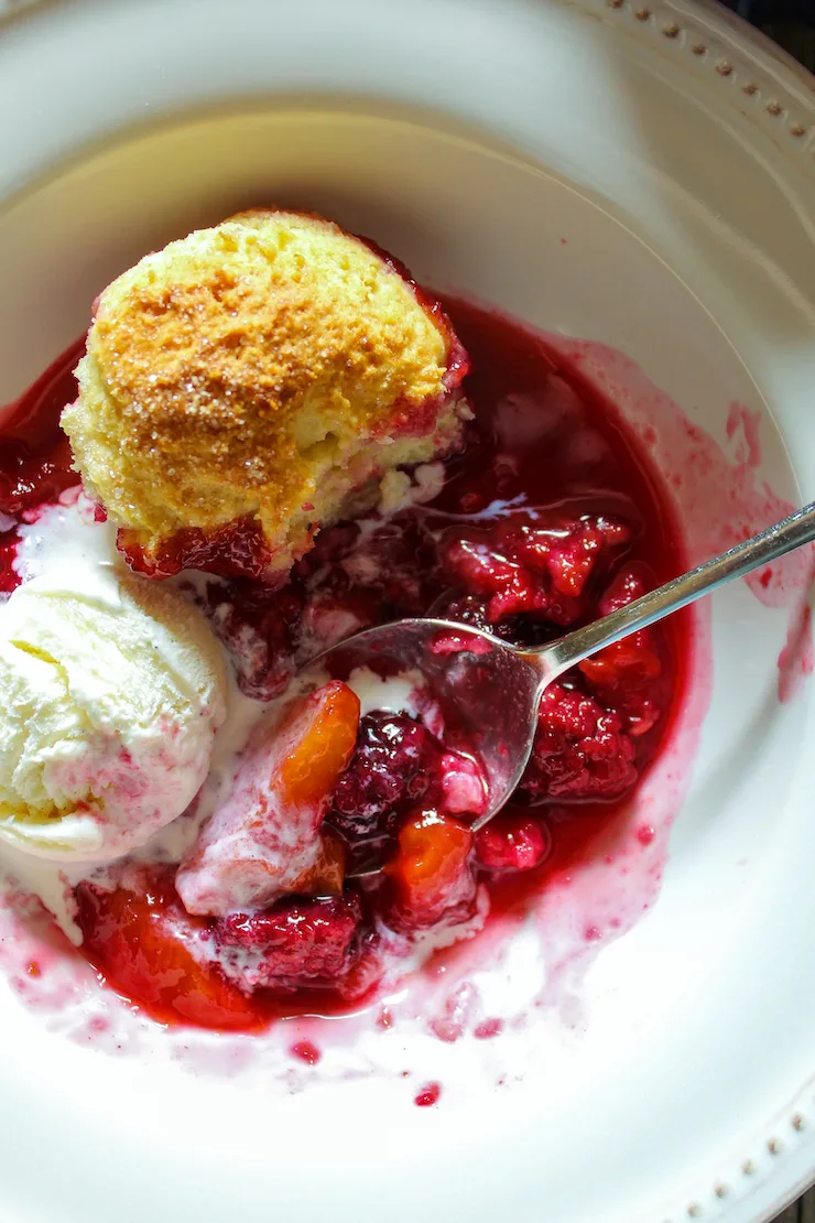 Closeup of blackberry peach cobbler with spoon and melting ice cream.