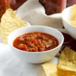 Salsa in bowl surrounded with chips and jars in background.