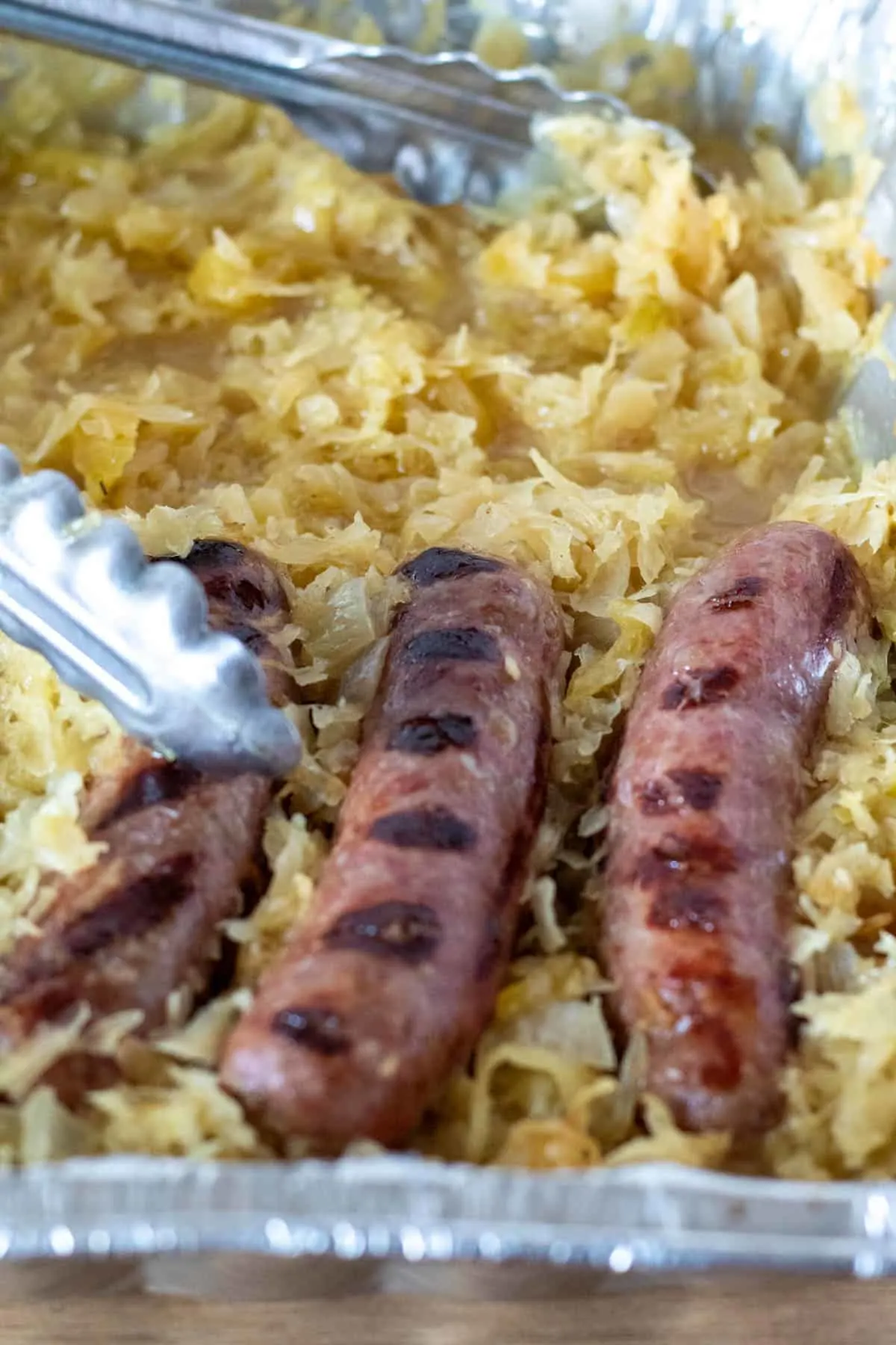 Grilled brats with tongs in disposable pan of sauerkraut.
