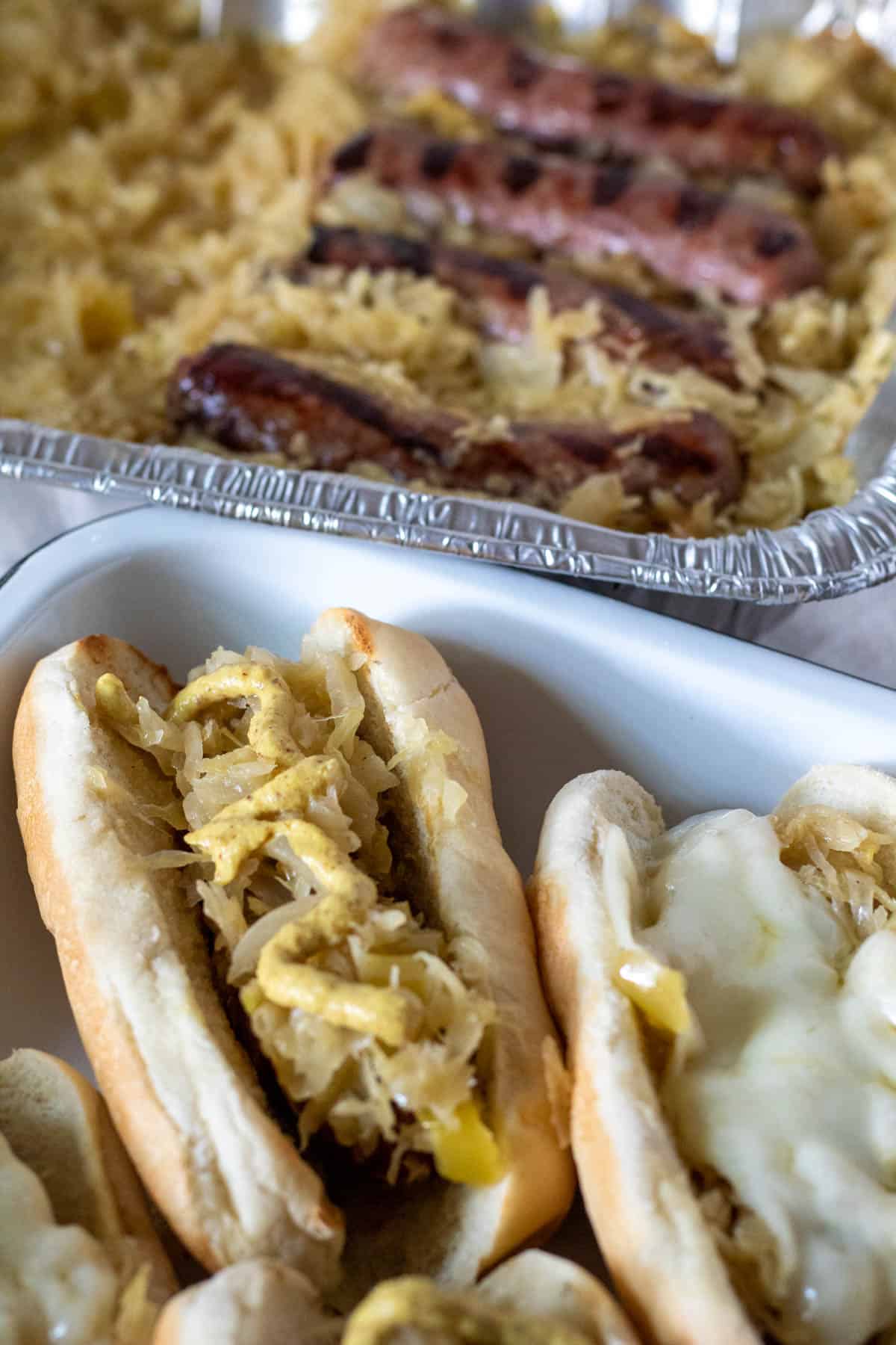 Brats on buns in pan with disposable pan in background.