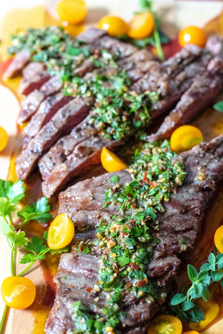 Sliced steak on cutting board with chimichurri and tomatoes.