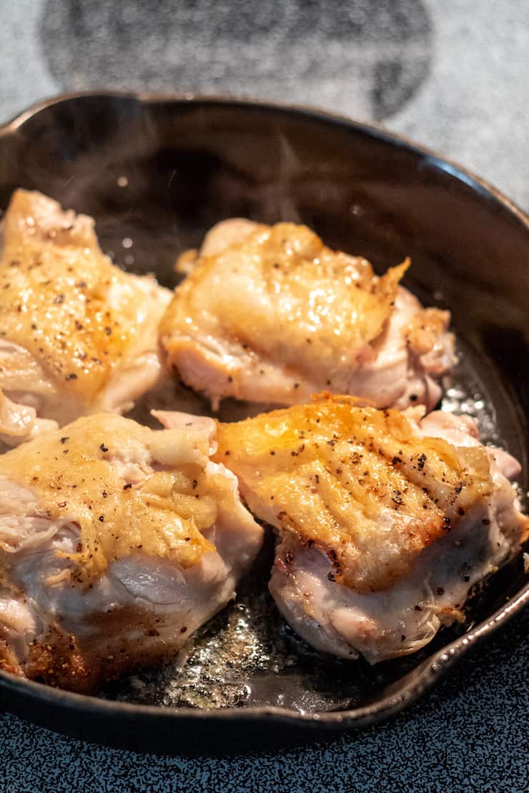 Chicken thighs browning on stovetop in cast iron skillet.