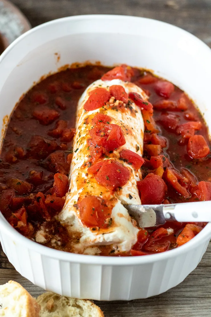 Goat cheese and tomatoes with serving knife in small casserole dish.