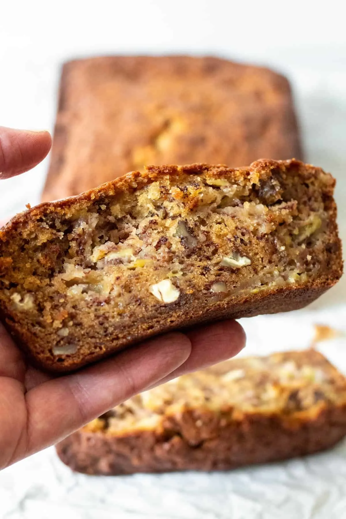 Hand holding a slice of banana bread with loaf in background.