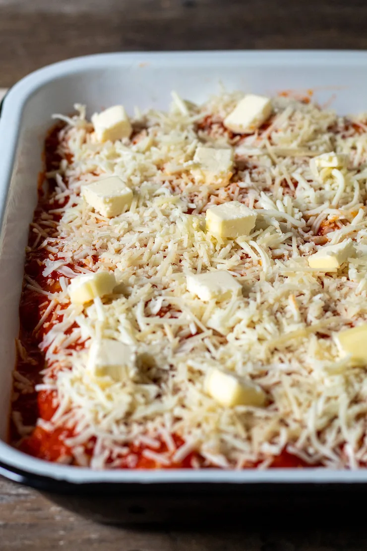 Assembled stuffed pasta tubes topped with shredded cheese and dotted with butter.