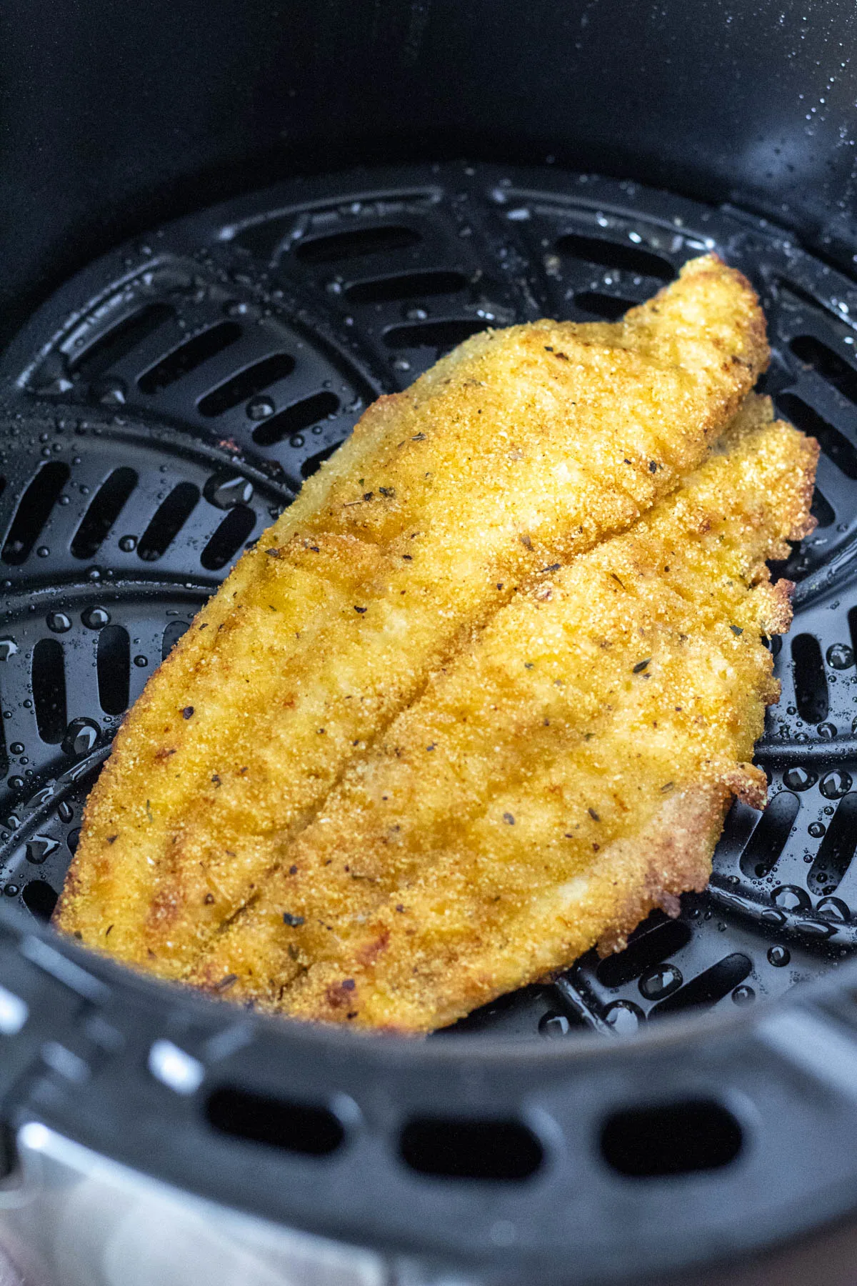 Catfish fillet in air fryer basket, browned on one side and ready to flip.