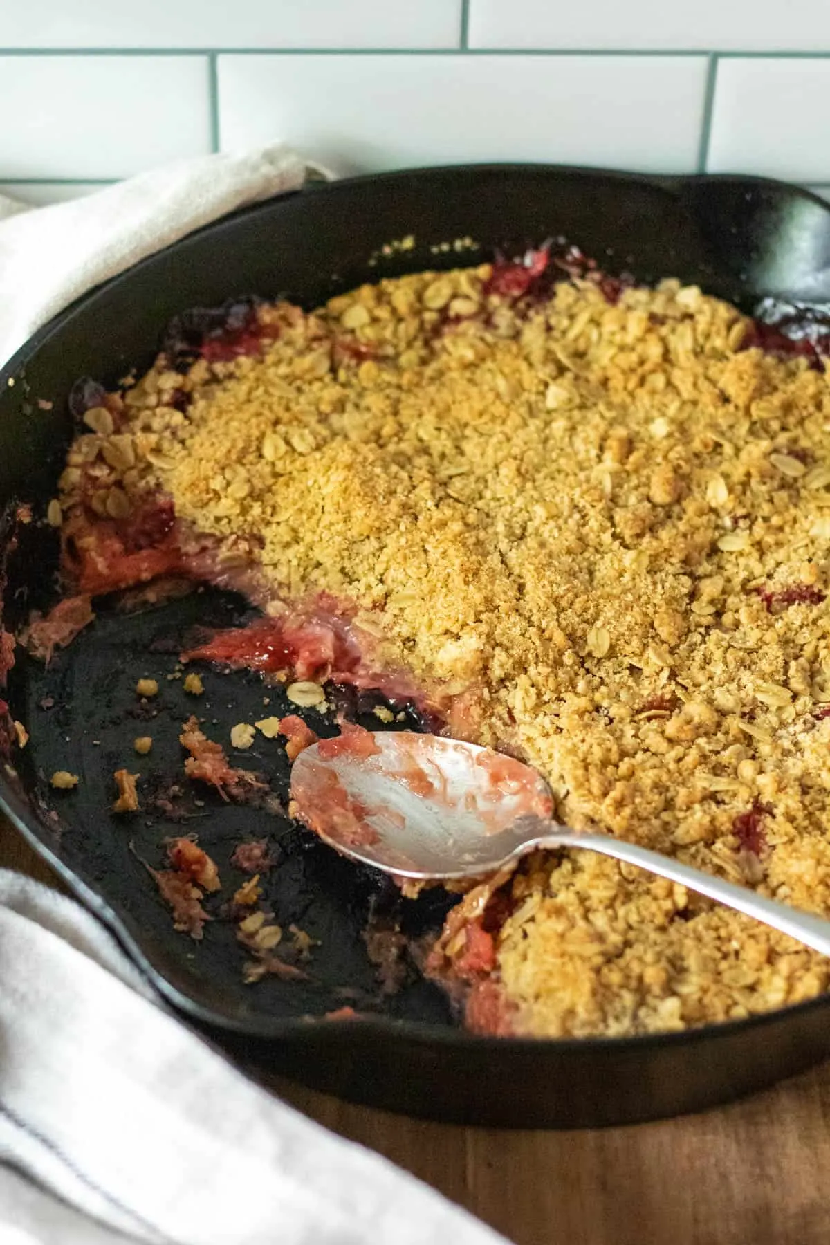 Strawberry rhubarb crisp in cast iron skillet, with serving spoon showing layers of crisp.