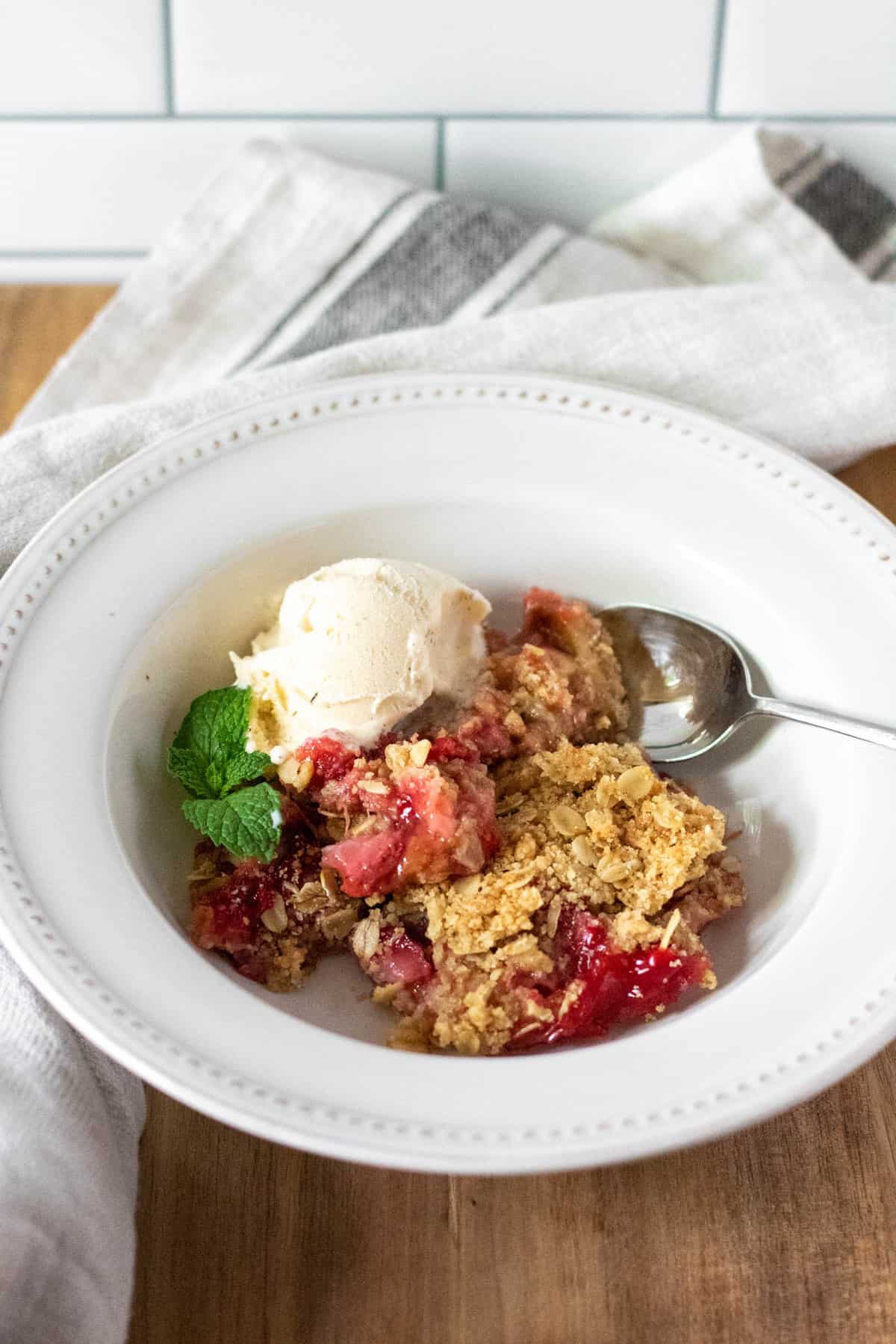 Strawberry rhubarb crisp in serving bowl with scoop of ice cream and spoon.