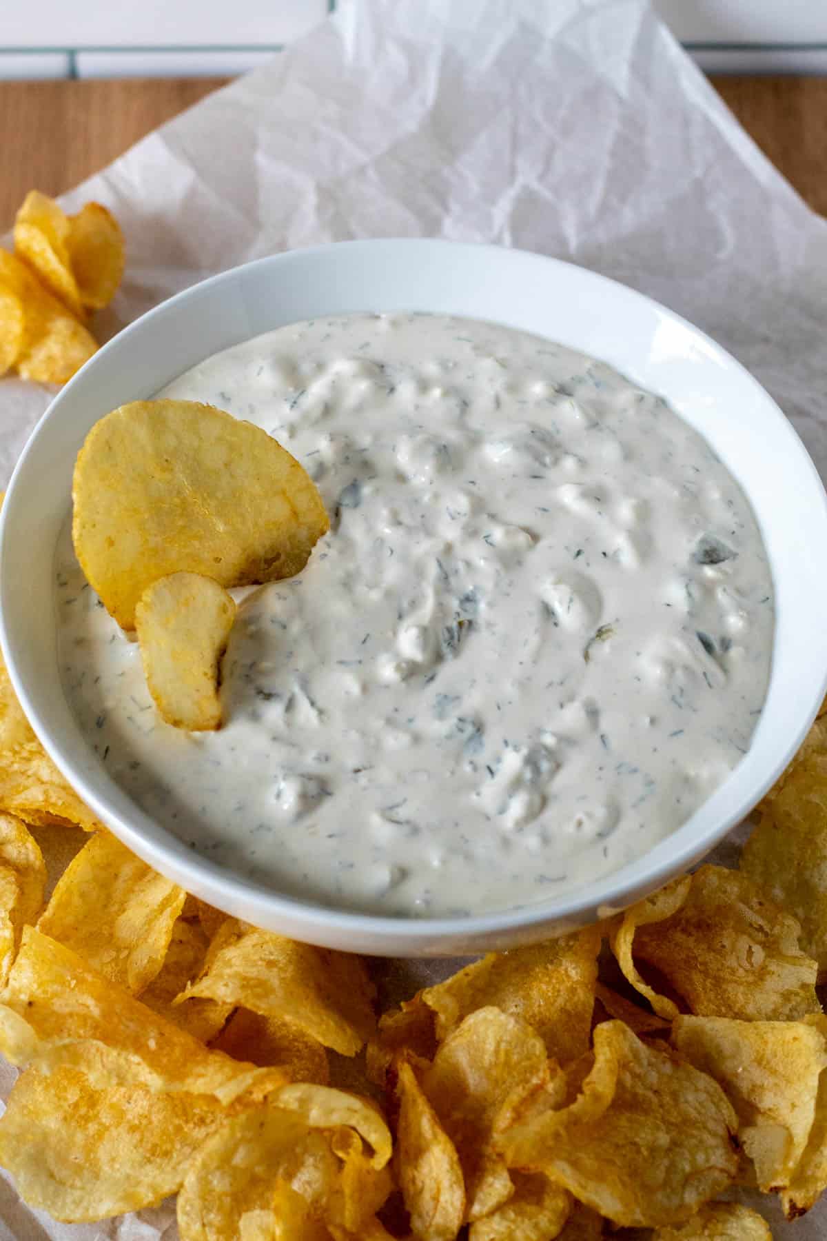Dill pickle dip in a serving bowl with two chips inseted on the side, bowl surrounded by potato chips.