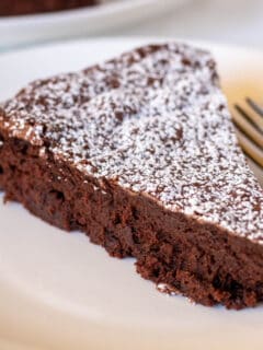 Piece of bittersweet chocolate cake with powdered sugar on white plate with fork.