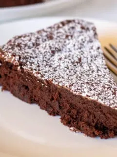 Piece of bittersweet chocolate cake with powdered sugar on white plate with fork.