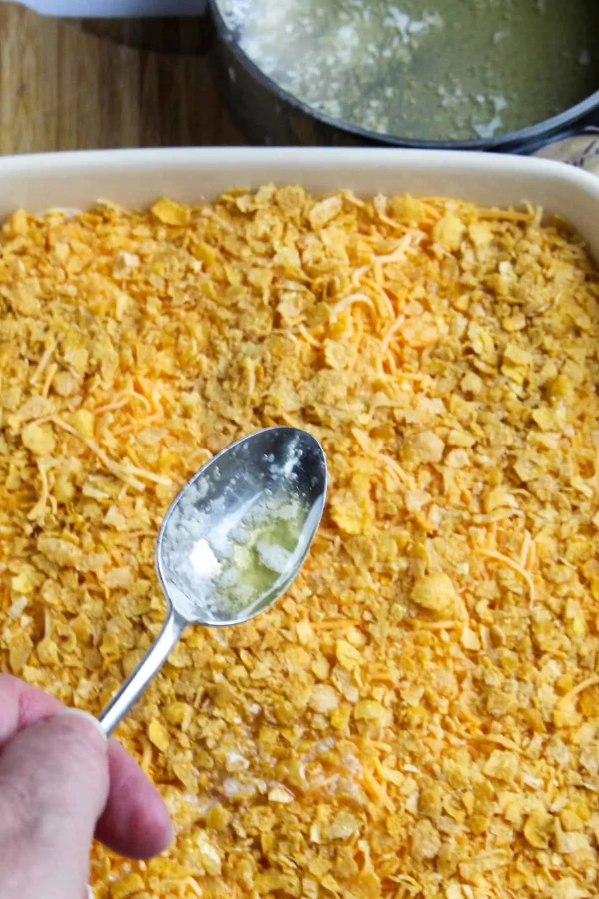 Finishing cheesy potato casserole with corn flakes and drizzling with melted butter over top with spoon.