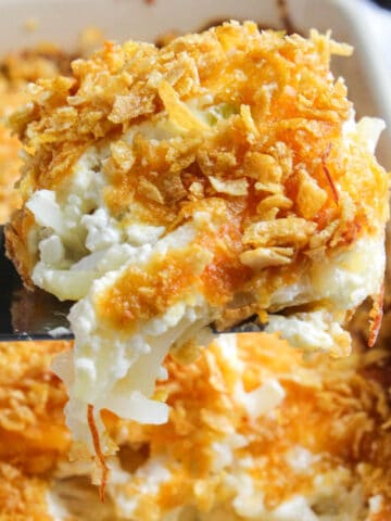 Close up of cheesy potatoes serving on spatula above casserole dish.